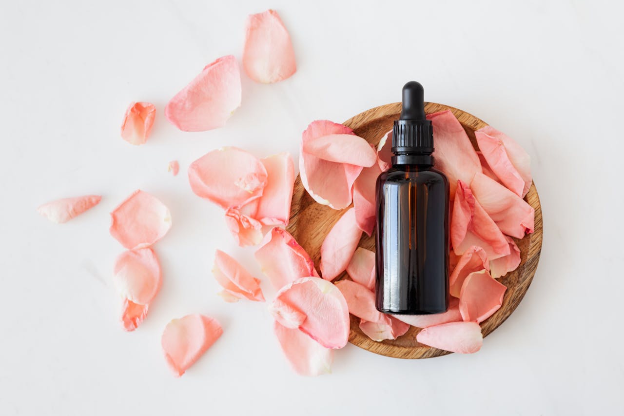 heros-img Top view of empty brown bottle for skin care product placed on wooden plate with fresh pink rose petals on white background isolated